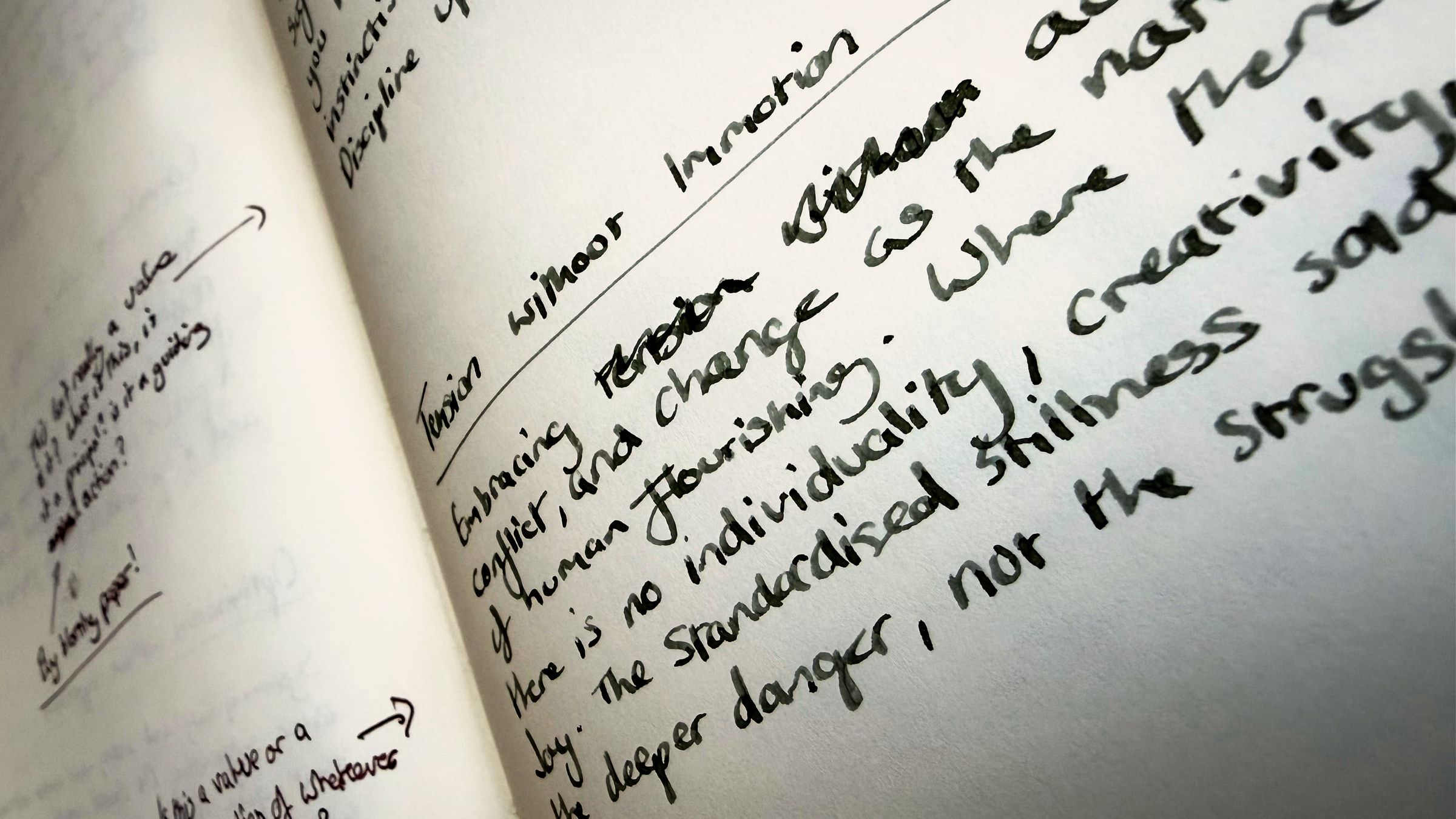 A notebook page photographed close and at an angle, the writing running away into soft focus. Sharp in the middle: the heading “Tension without Immotion” and the start of the argument this part makes, that active tension, conflict, and change are the natural condition of human flourishing, and that the standardised stillness sold as peace is the deeper danger. Two words are struck through and rewritten mid-sentence. On the facing page, out of focus, are later notes in a second pen.
