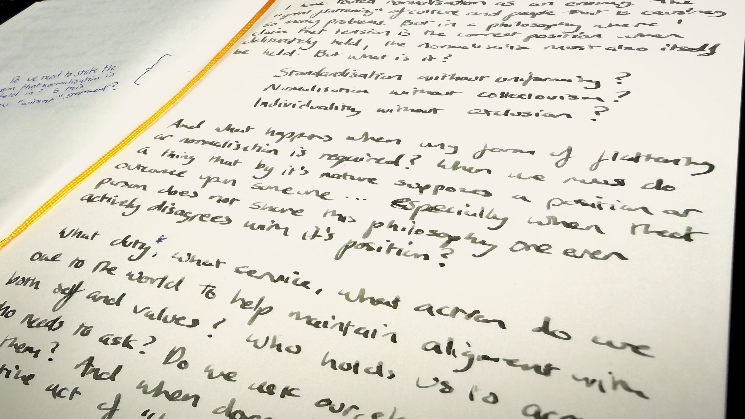 A notebook page photographed close and at an angle, the writing running away into soft focus. Sharp across the middle, the question this piece asks: “what duty, what service, what action do we owe to the world to help maintain alignment with both self and values? Who holds us to account? Who needs to ask?” Above it, out of focus, three more: standardisation without uniformity, normalisation without collectivism, individuality without exclusion. The notebook’s elastic runs down the gutter.